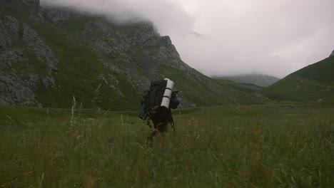Man-is-hiking-in-a-valley,-moving-through-grass-and-having-mountains-with-fog-in-front-of-him