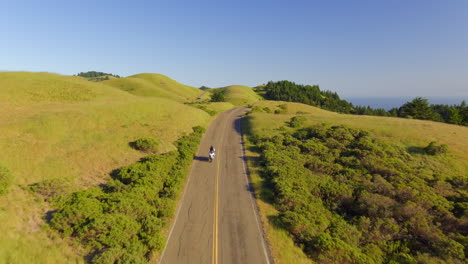 A-motorcycle-driving-along-a-picturesque-countryside-road-along-green,-grassy-hills-near-Mt-Tamalpais,-California---aerial-flyover