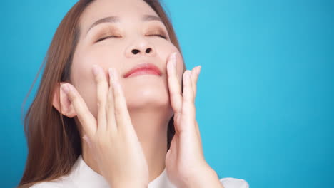 Nahaufnahme-Einer-Perfekten-Hübschen-Asiatischen-Frau,-Die-Das-Gesicht-Sanft-Mit-Anti-Aging-Balsamcreme-Für-Wellness-Hautpflege-Auf-Blauem-Hintergrund-Reibt-2