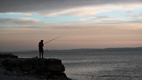 Old-man-catching-a-fish-from-a-rocky-pier-during-beautiful-summer-sunset