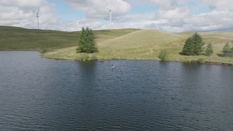 Establishing-Aerial-Shot-Of-A-Young-Woman-On-A-Stand-Up-Paddleboard-With-Windmills-Spinning-In-The-Background