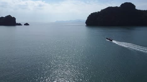 Tourist-boat-in-blue-ocean-with-silhouetted-rocks-in-Langkawi-Malaysia,-drone-view