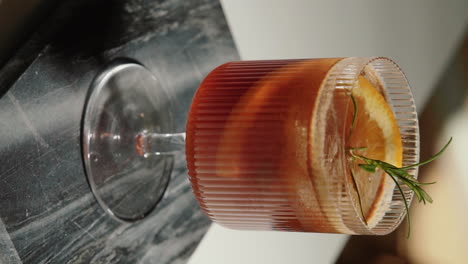 Vertical-Video-Presenting-an-Espresso-tonic-with-cold-brew-coffee,-orange-slice-and-rosemary-branch-in-the-kitchen-at-home