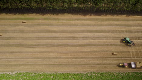 Aerial-top-down-shot-of-tractor-loading-harvested-hay-bales-on-tractor-trailer-at-farm-field