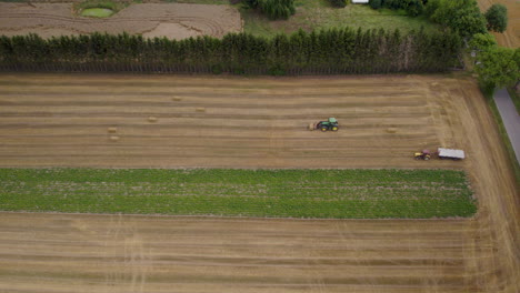 Industrial-tractor-picking-hay-bales-from-agricultural-field-and-driving-backwards---aerial-top-down-shot