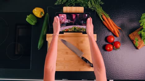 Birds-eye-shot-of-a-women-put-tablet-on-cutting-board-on-a-with-a-recipe-on-a-tablet-in-a-modern-kitchen-and-fresh-herbs-and-vegetables