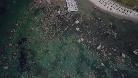 Vista-De-Drones-Sobre-Las-Olas-Del-Mar-Que-Revelan-Una-Playa-Rocosa-Y-Sombrillas-De-Paja-Cerca-Del-Bar