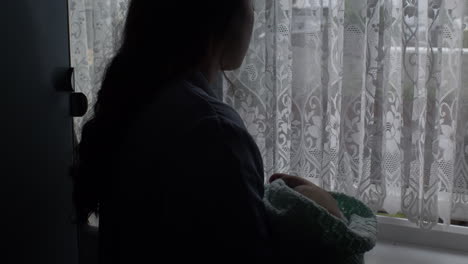 Silhouette-Of-Young-Mother-Cradling-Her-Newborn-Baby-In-Her-Arms-Beside-Windowsill-And-Gently-Swaying-Side-To-Side-Locked-Off