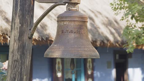 Hundred-years-old-copper-bell-in-front-of-traditional-wooden-hut-with-thatched-roof