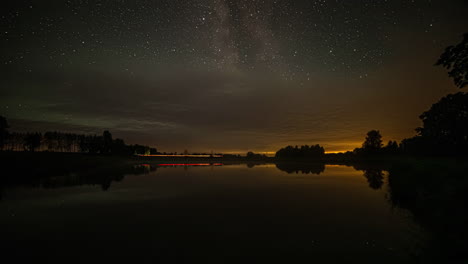 Timelapse-shot-of-stars-visible-just-before-sunrise-over-a-road-beside-a-lake-in-rural-countryside