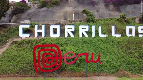 Drone-video-of-a-red-sign-reading-"Peru"-and-behind-it-on-a-green-hill,-another-sign-reading-"Chorrillos"-the-name-of-the-district-in-Lima,Peru
