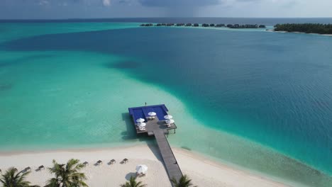 Aerial-view-of-luxury-resort-in-Maldives-with-palm-trees-on-beach-and-overwater-infinity-pool-with-bungalows-in-background