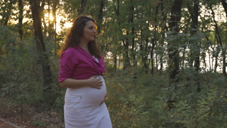 Pregnant-Woman-Expectant-Mother-Walking-Alone-in-Park-Forest-at-Sunset,-Attractive-Young-Casual-Dressed-Mom-Stroking-Caressing-Touching-Her-Belly-with-Hands-Smiling,-Medium-Portrait-Tracking-Shot