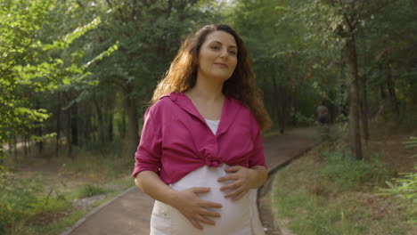 Happy-Pregnant-Woman-Expectant-Mother-Walking-Alone-in-Park-Turning-Around,-Attractive-Young-Casual-Dressed-Mom-Stroking-Caressing-Touching-Her-Belly-with-Hands-Smiling,-Medium-Portrait-Shot