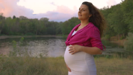 Portrait-of-Pregnant-Woman-Expectant-Mother-Walking-Alone-in-Forest-Next-to-Lake,-Attractive-Caucasian-Casual-Dressed-Mom-Stroking-Caress-Touching-Her-Belly-with-Hands-Smiling,-Childbearing-Tenderness