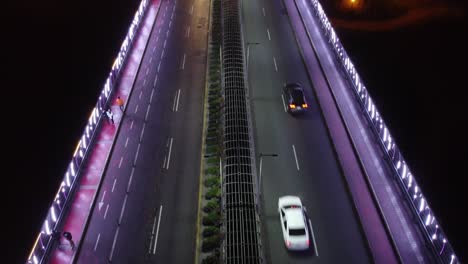 Drone-video-of-a-bridge-at-night-lit-with-purple-lights,-People-walk-by-on-sidewalk-as-cars-drive-by-in-the-street-lanes