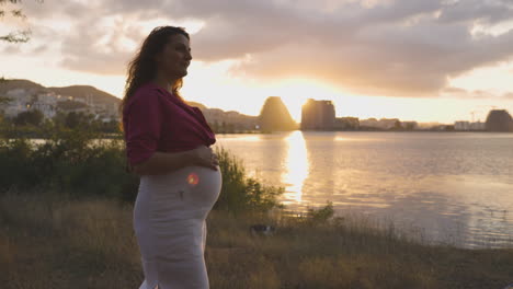 Mujer-Embarazada-Futura-Madre-Caminando-Sola-Por-El-Lago-Al-Atardecer,-Atractiva-Joven-Madre-Vestida-Casualmente-Acariciando-Acariciando-Su-Vientre-Con-Las-Manos-Sonriendo,-Retrato-De-Muñeca-Mediana-Embarazada