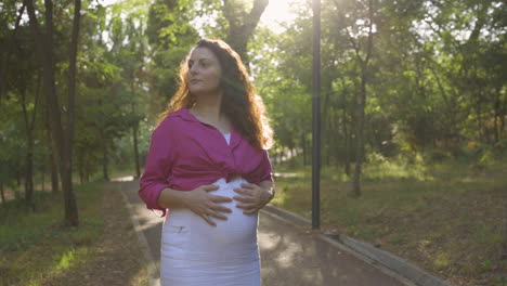 Pregnant-Woman-Expectant-Mother-Walking-Alone-in-Park,-Attractive-Caucasian-Casual-Dressed-Mom-Stroking-Caressing-Touching-Her-Belly-with-Hands-Relaxing,-Childbearing-Tenderness,-Medium-Portrait-Shot