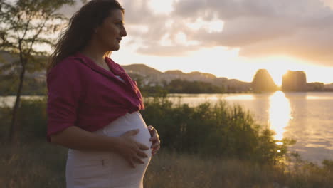 Schwangere-Frau-Werdende-Mutter,-Die-Bei-Sonnenuntergang-Allein-Am-See-Spazieren-Geht,-Attraktive-Kaukasische,-Lässig-Gekleidete-Mutter,-Die-Ihren-Bauch-Mit-Lächelnden-Händen-Berührt,-Gebärfähige-Mittlere-Porträtaufnahme