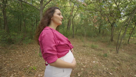 Tracking-Shot-of-Pregnant-Woman-Expectant-Mother-Walking-Alone-in-Forest,-Attractive-Caucasian-Casual-Dressed-Mom-Stroking-Caress-Touching-Her-Belly-with-Hands-and-Smiling,-Childbearing-and-Tenderness