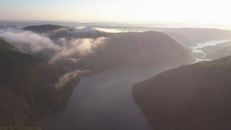 Wide,-high-altitude-aerial-view-of-Tarnita-Lake,-Romania-as-day-breaks-with-golden-light-on-landscape-with-light-fog-and-soft-low-white-clouds