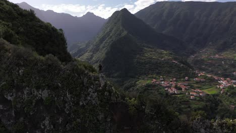 Kreisende-Drohnenaufnahme-Eines-Mannes,-Der-In-Einer-Kraftvollen-Und-Selbstbewussten-Heldenpose-Am-Rande-Des-Berges-Steht-Und-Die-Wunderschöne-Landschaft-Madeiras-überblickt