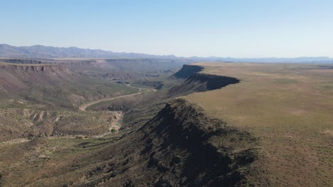 Drone-Volando-Sobre-El-Borde-Exterior-De-Un-Cañón-En-El-Desierto-Alto-En-Arizona,-Cielo-Azul-Y-Montañas-En-El-Fondo