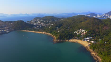 Drone-shot-traveling-to-the-left-next-to-a-urbanized-tropical-island-with-some-villages-and-beach-among-the-mountains-and-forest-during-a-sunny-day