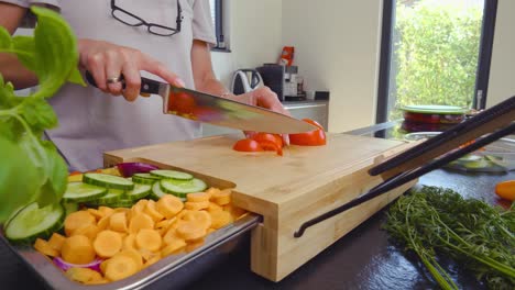 Panning-reveal-shot-of-a-person-cutting-a-tomato-on-a-wood-cutting-board-inside-a-modern-style-kitchen-with-cut-vegetables-in-preparation-for-a-meal