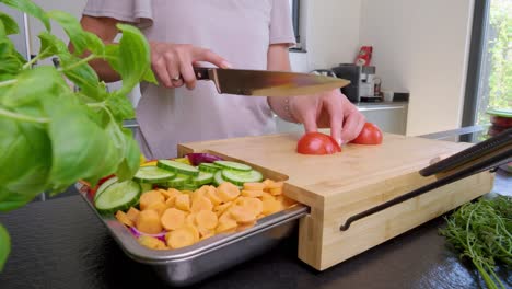 Panning-Enthüllt-Eine-Aufnahme-Einer-Person,-Die-Eine-Tomate-Auf-Einem-Holzschneidebrett-In-Einer-Modernen,-Stilvollen-Küche-Mit-Einem-Tablett-Mit-Zubereitetem-Gemüse-Schneidet