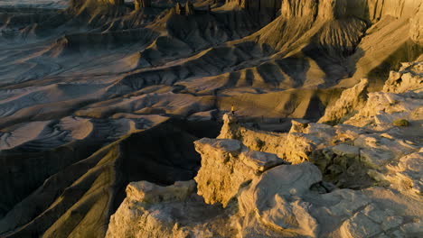 Tapfere-Frau-In-Gelber-Jacke,-Die-Am-Rand-Einer-Steilen-Felsklippe-Mit-Blick-Auf-Die-Karge-Landschaft-Bei-Einem-Sonnenaufgang-In-Utah-Steht