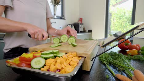 Orbiting-shot-of-a-person-cutting-cucumbers-on-a-cutting-board-with-other-fresh-vegetables-prepared,-in-a-modern-clean-home-kitchen-on-a-bright-sunny-day