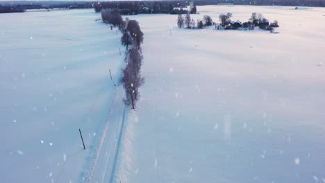 Lonely-tractor-transporting-haybales-on-icy-winter-road-during-snowfall,-aerial-view