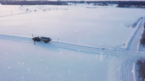 Tractor-transporting-haybales-during-heavy-snowfall-in-winter-season,-aerial-view