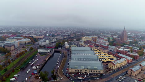 Vuelo-Aéreo-Hacia-Atrás-Sobre-La-Ciudad-De-Riga-Con-Market-Hall-Durante-El-Día-Nublado,-Letonia