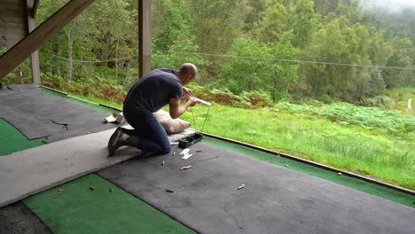 Man-at-shooting-range-assembles-ballistic-precision-chronograph-for-testing-bullet-speed---Empty-brass-shells-on-ground-and-green-forest-background