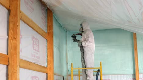 Man-in-disposable-Tyvek-suit-and-respirator-is-spraying-foam-insulation-near-the-ceiling-of-exterior-wall-while-standing-on-scaffolding