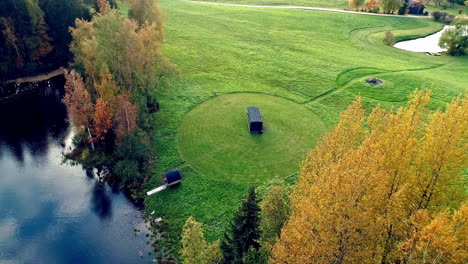 Aerial-shot-of-grass-field-with-barrel-sauna-next-to-natural-lake-during-daytime