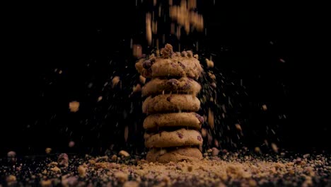 Stack-of-vegan-chocolate-cookies-with-biscuit-crumbs-falling-on-them-while-they-rotate,-slow-motion,-black-background,-static-shot