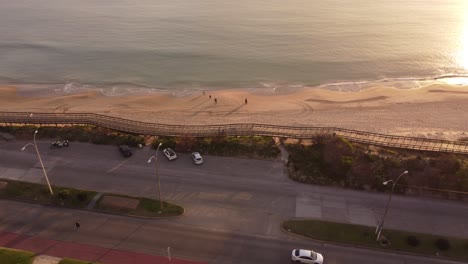 Aerial-view-of-people-walking-along-sandy-beach-and-coast-of-Punta-del-Este-and-enjoying-golden-sunset---Cars-driving-along-coastal-road-in-the-evening