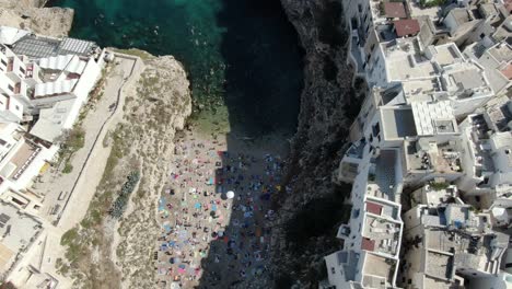 Drone-Captura-La-Concurrida-Playa-De-Polignano-A-Mare-Con-Numerosas-Personas-Descansando-Bajo-La-Sombrilla,-Algunos-Pasean-Por-La-Playa-Y-Otros-Nadan-En-El-Mar