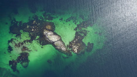 Dramatic-aerial-panning-birdseye-view-of-rocky-island-with-green-ocean,-waves,-sandy-bottom-and-sun-glare-on-the-ripples-of-the-water