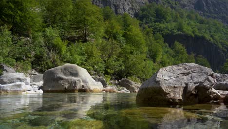Tranquil-river-water-surface-reflecting-big-cliffs-and-green-trees-on-Alpine-mountains