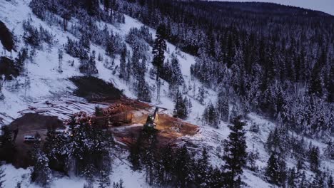 Drone-dramatically-moves-towards-timber-manipulator-tractor-sorting-through-snow-covered-piles-of-cut-mature-pine-trees-in-timber-logging-camp-as-loaded-18-wheeler-pulls-away