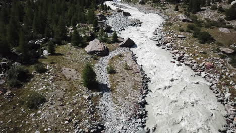 Drone-view,-tilt-up-of-a-rocky-and-muddy-river-from-the-Swiss-alps-with-strong-current,-mountain-valley