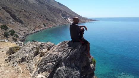 Barefoot-meditating-man-sitting-on-the-edge-of-a-cliff-contemplating-the-panoramic-view-of-the-amphitheater-bay-of-Loutro