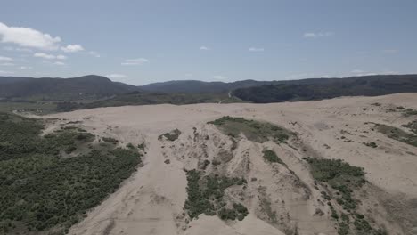 Push-in-aerial-shot-of-sand-dunes-in-Llani,-Chile-with-bushes-on-the-sides-and-mountains-in-the-background-taken-during-a-clear-sunny-day-with-some-clouds