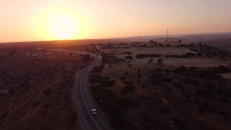 Aerial-drone-footage-of-a-scenic-rural-street-with-a-white-pick-up-car-passing-leading-to-the-sunset-from-above