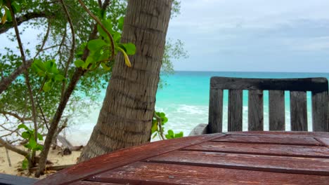 Rustic-Wood-Table-And-Chair-Beside-Tropical-Beach-And-Tree-With-Turquoise-Waves-Breaking-In-Background