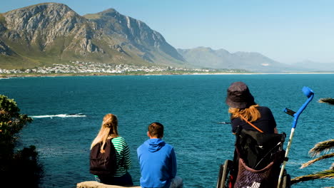 Lady-in-wheelchair-on-Hermanus-coastline-at-vantage-point-doing-land-based-whale-watching---panoramic-view-over-bay-toward-mountains